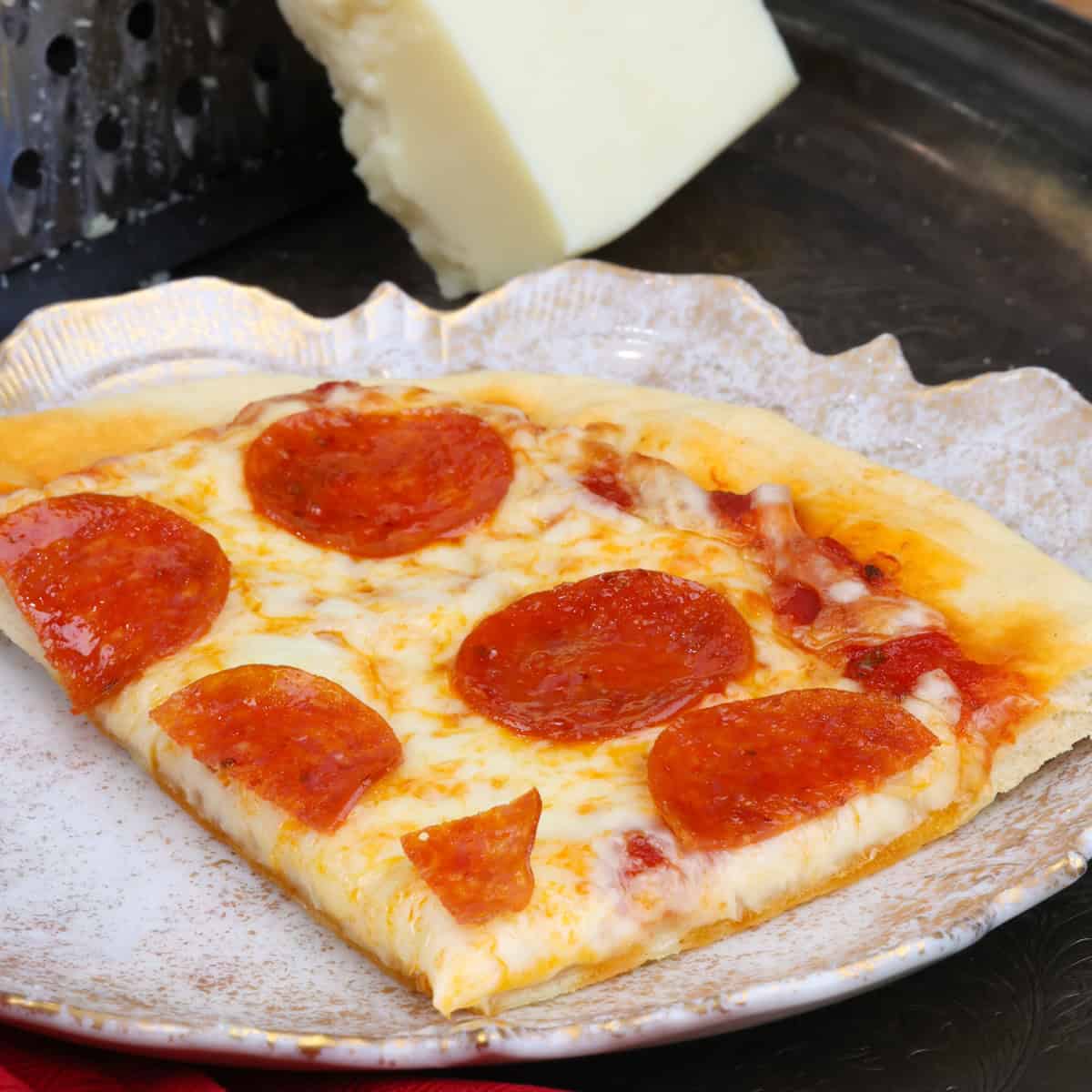 a slice of pepperoni pizza on a white plate next to a block of mozzarella cheese and a red napkin.
