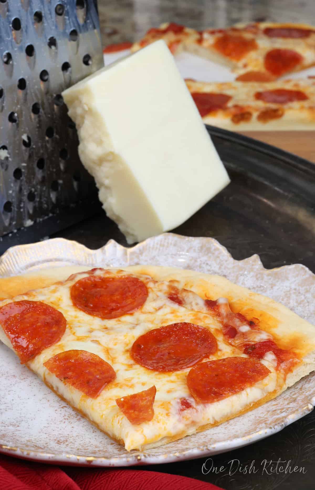 a slice of pepperoni pizza on a white plate next to a block of mozzarella cheese and a red napkin.