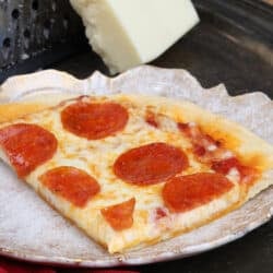 a slice of pepperoni pizza on a white plate next to a block of mozzarella cheese and a red napkin