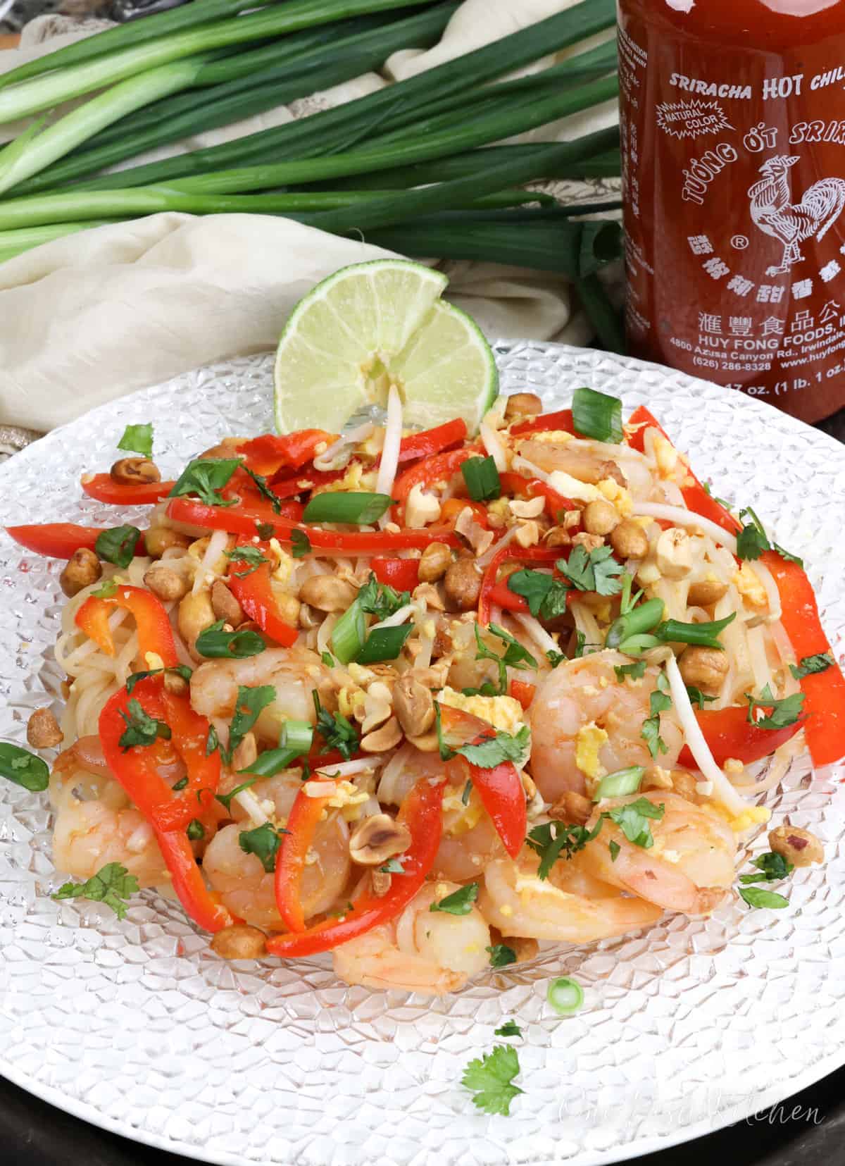 pad thai on a white plate next to two lime wedges, a bottle of sriracha, and several green onions