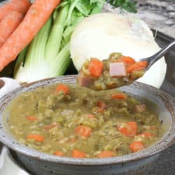 a spoonful of split pea soup over a grey bowl next to an onion, celery, and carrots.