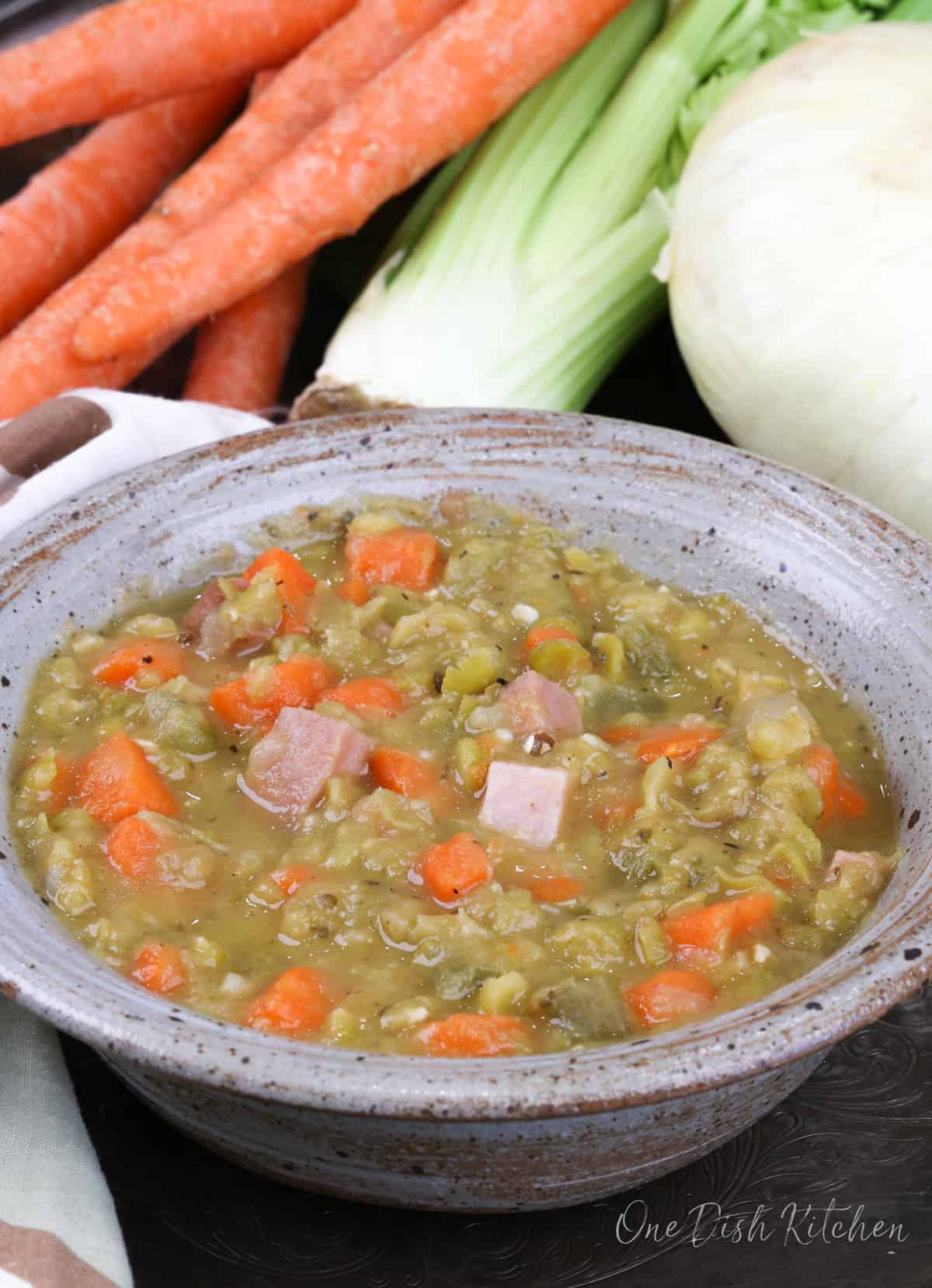 a blue bowl filled with split pea soup next to an onion, celery, and several carrots on a silver tray