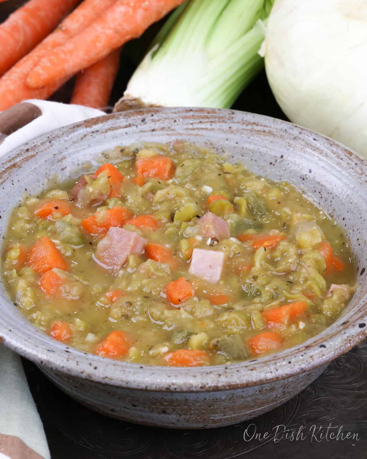 a blue bowl filled with split pea soup next to an onion, celery, and several carrots on a silver tray.