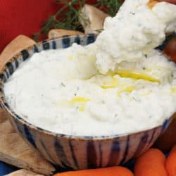 tzatziki sauce topped with olive oil surrounded by pita bread, carrots, and a red napkin and being scooped out with pita chip