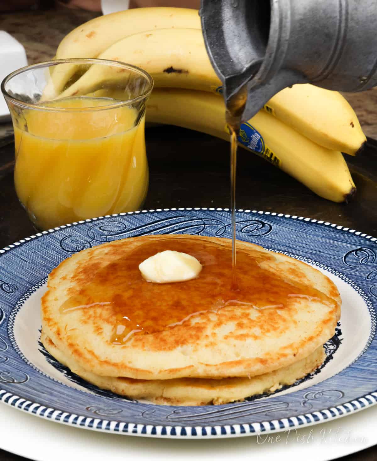 two buttermilk pancakes on a blue plate topped with butter and syrup next to a glass of orange juice and bananas.