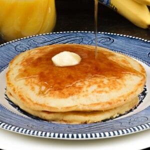 two buttermilk pancakes on a blue plate topped with butter and syrup next to a glass of orange juice and bananas
