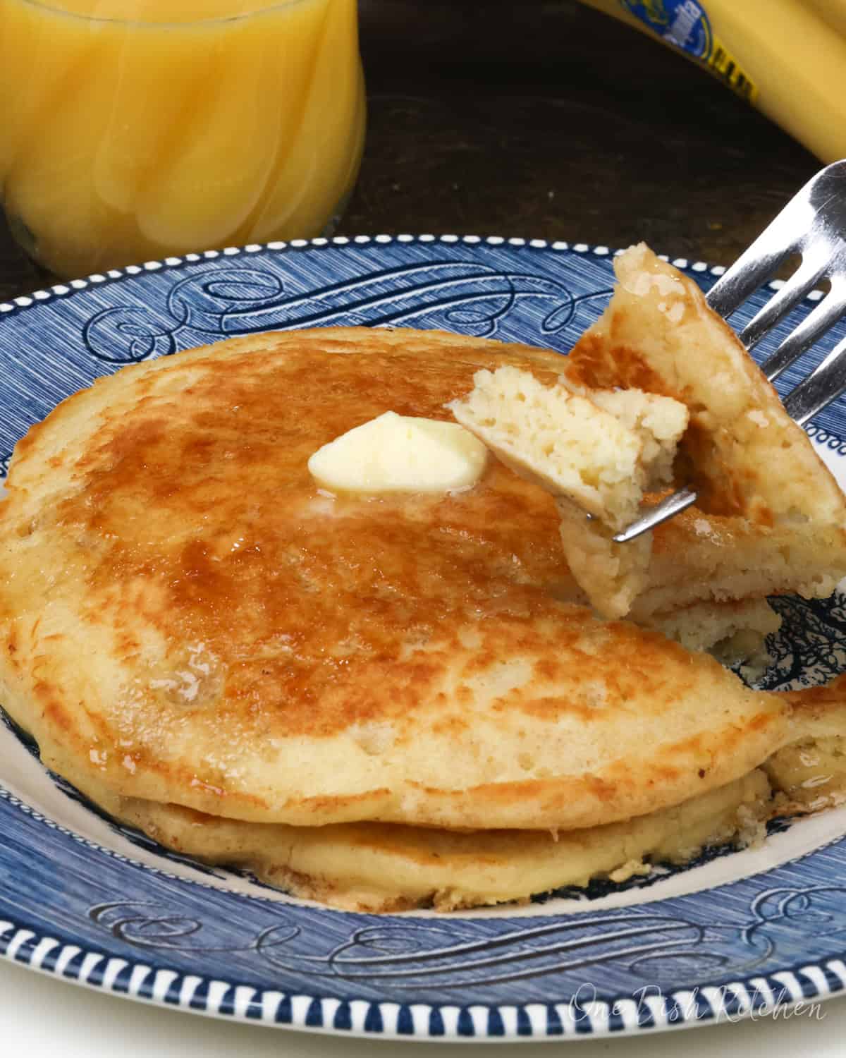 two buttermilk pancakes on a blue plate topped with butter and syrup next to a glass of orange juice and bananas.