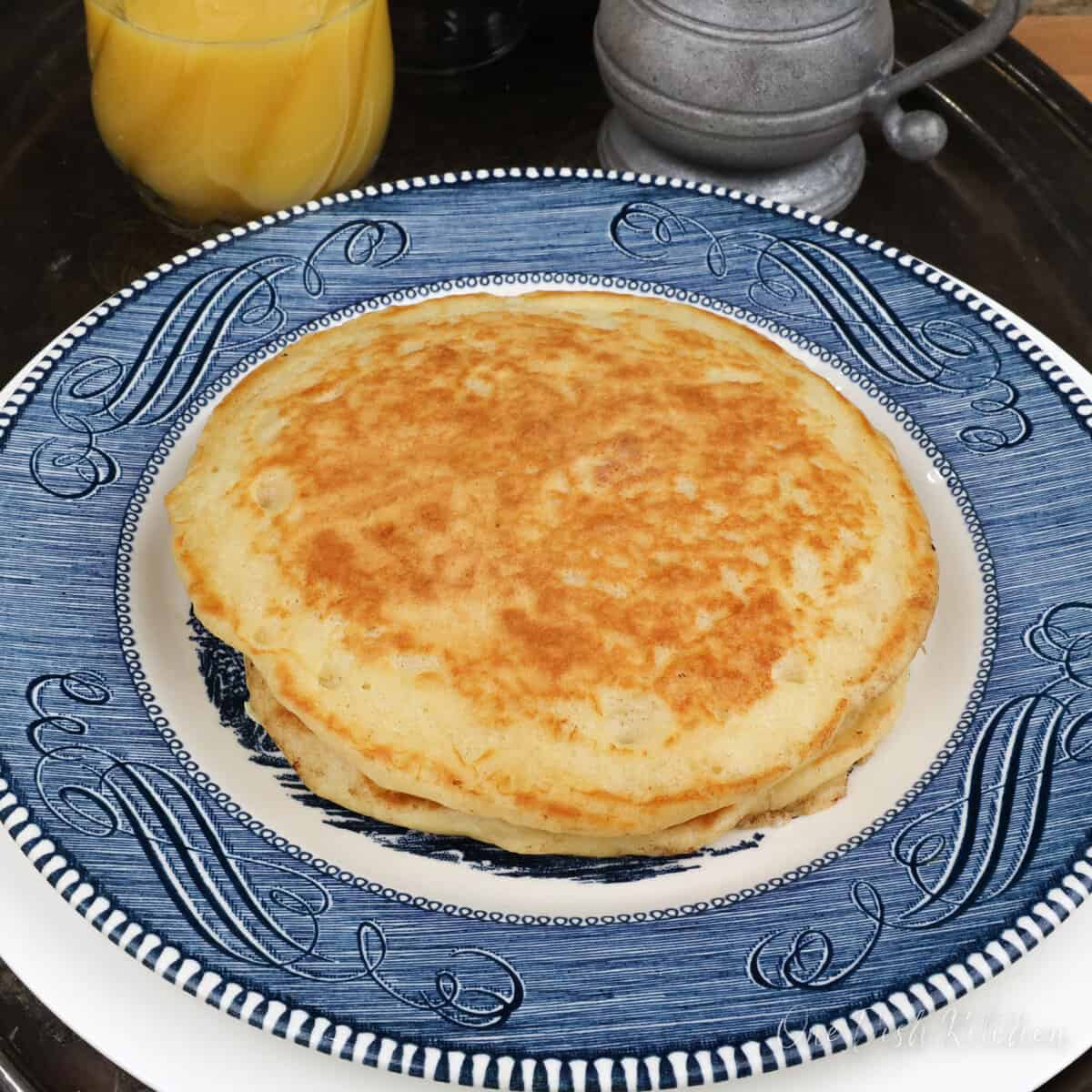 two buttermilk pancakes on a blue plate next to a glass of orange juice and a silver container of syrup