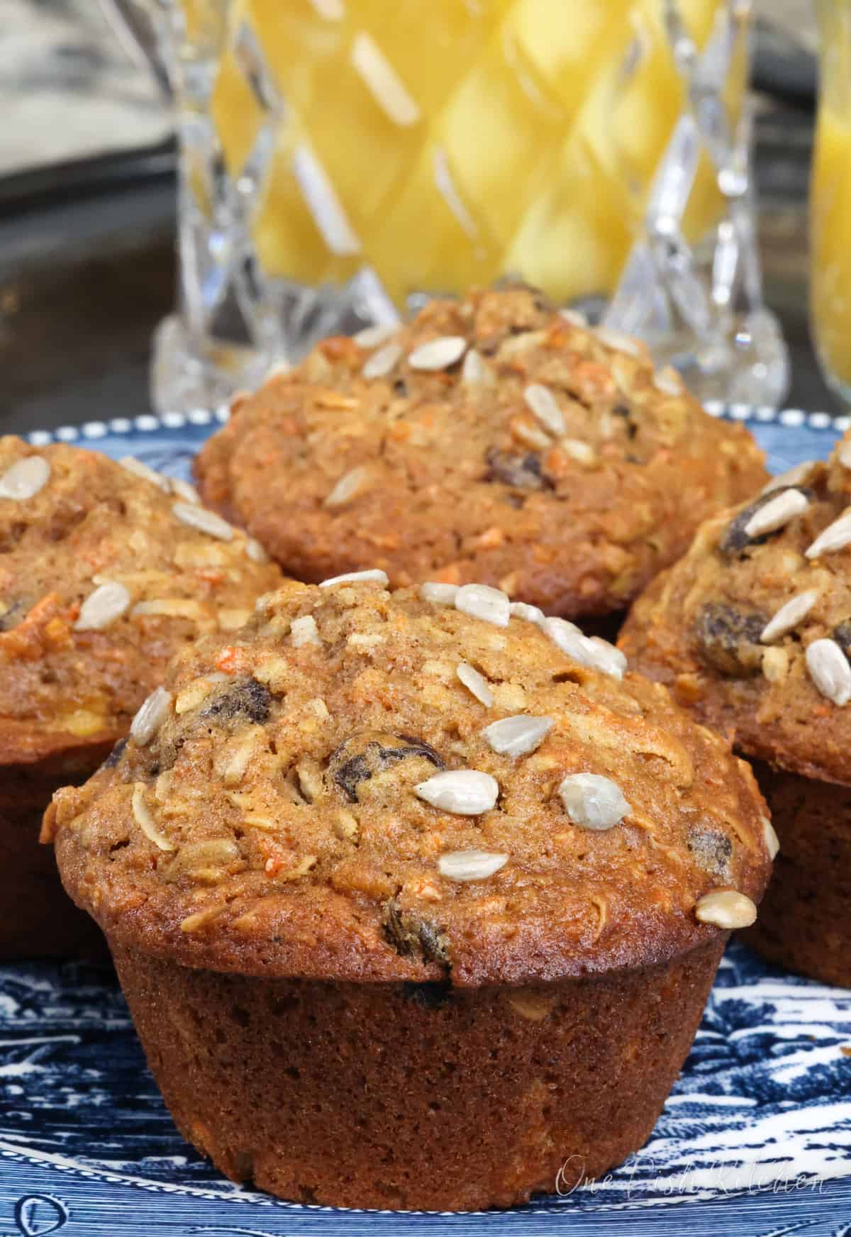 four morning glory muffins on a blue plate next to a pitcher of orange juice.
