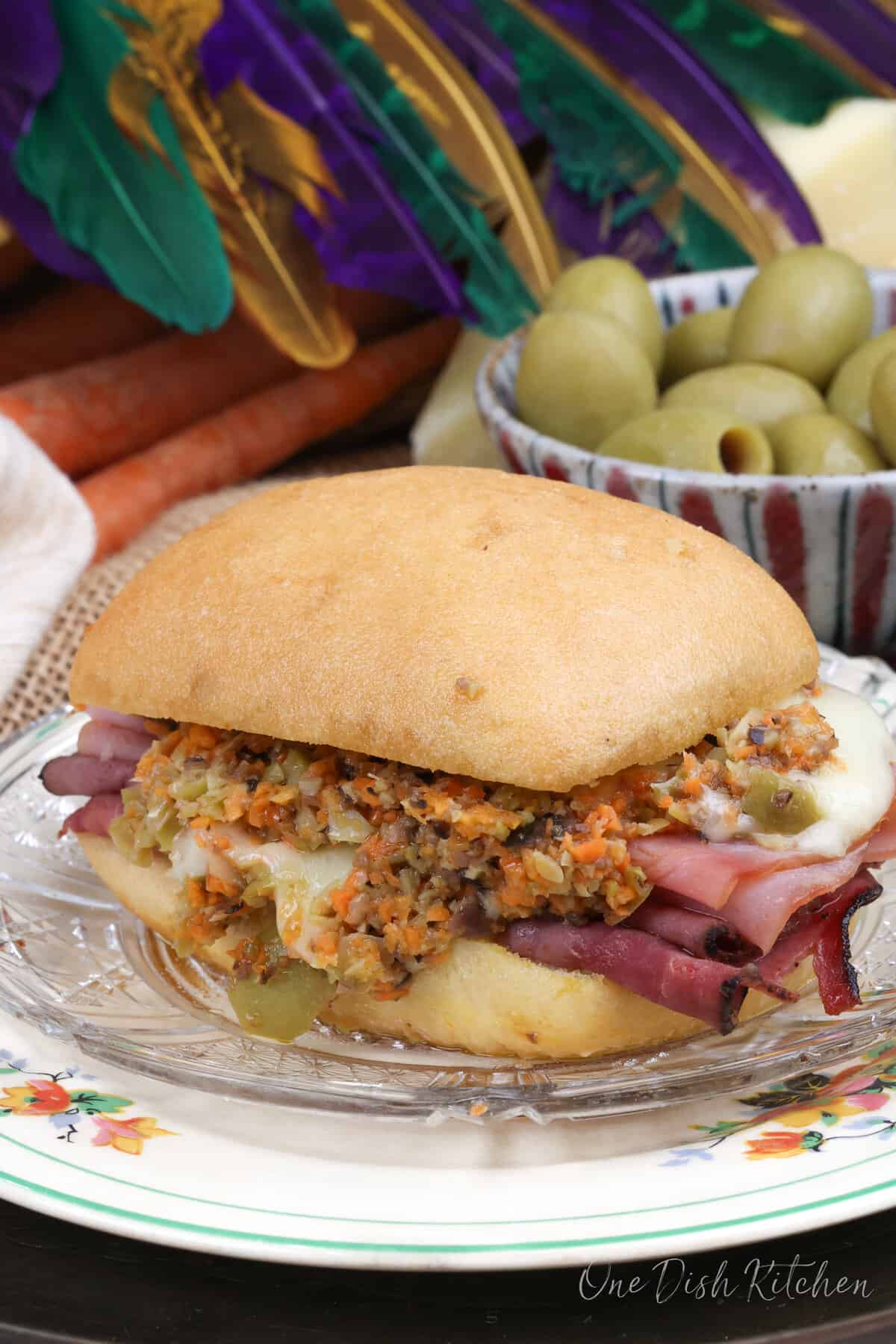 a muffuletta on a white plate next to a purple green and gold mardi gras mask and a bowl of green olives.