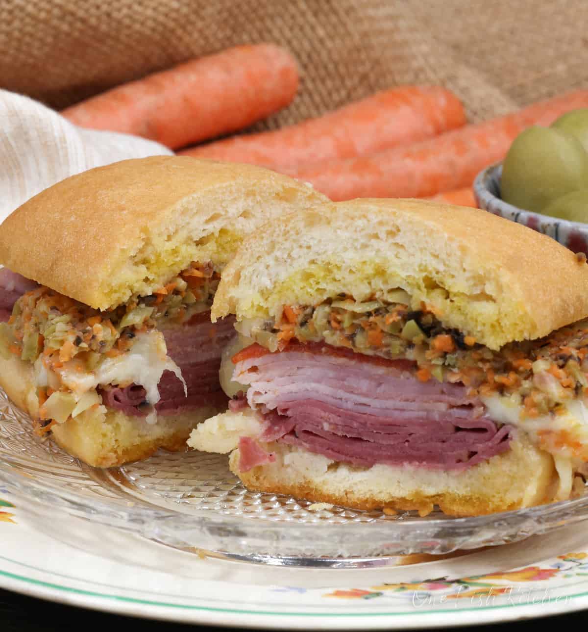 a muffuletta sliced in half next to 3 carrots and a bowl of olives.
