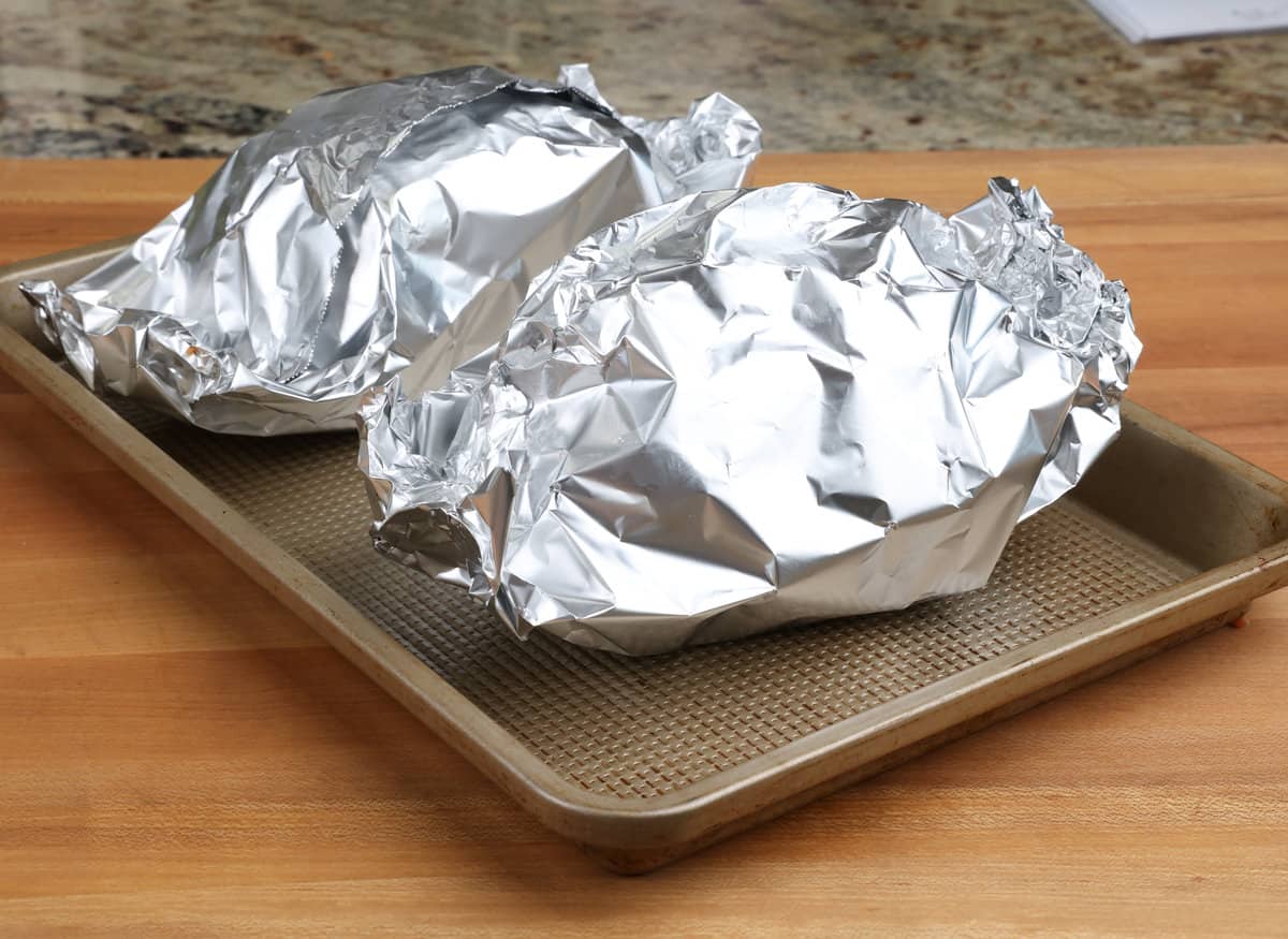 two mini muffulettas wrapped in aluminum foil on a baking sheet.