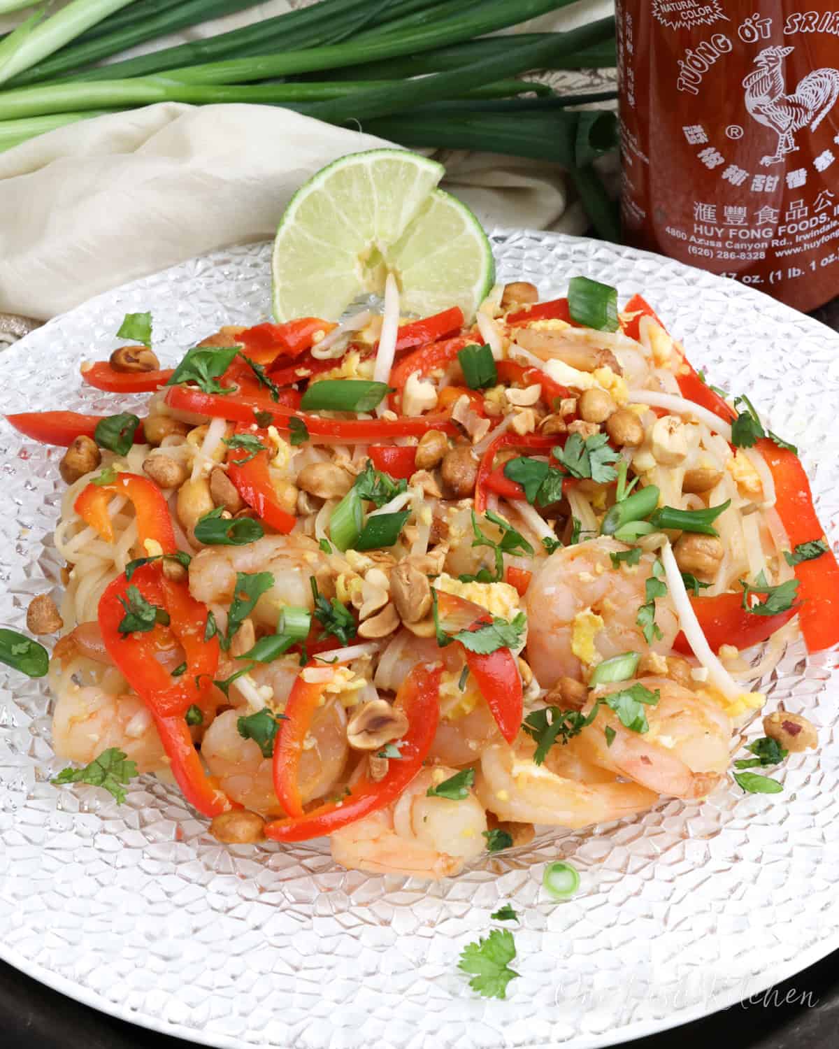 pad thai on a white plate next to two lime wedges, a bottle of sriracha, and several green onions.