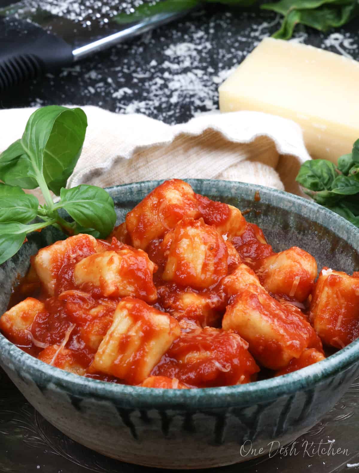 potato gnocchi topped with tomato sauce in a green bowl next to fresh basil and parmesan cheese
