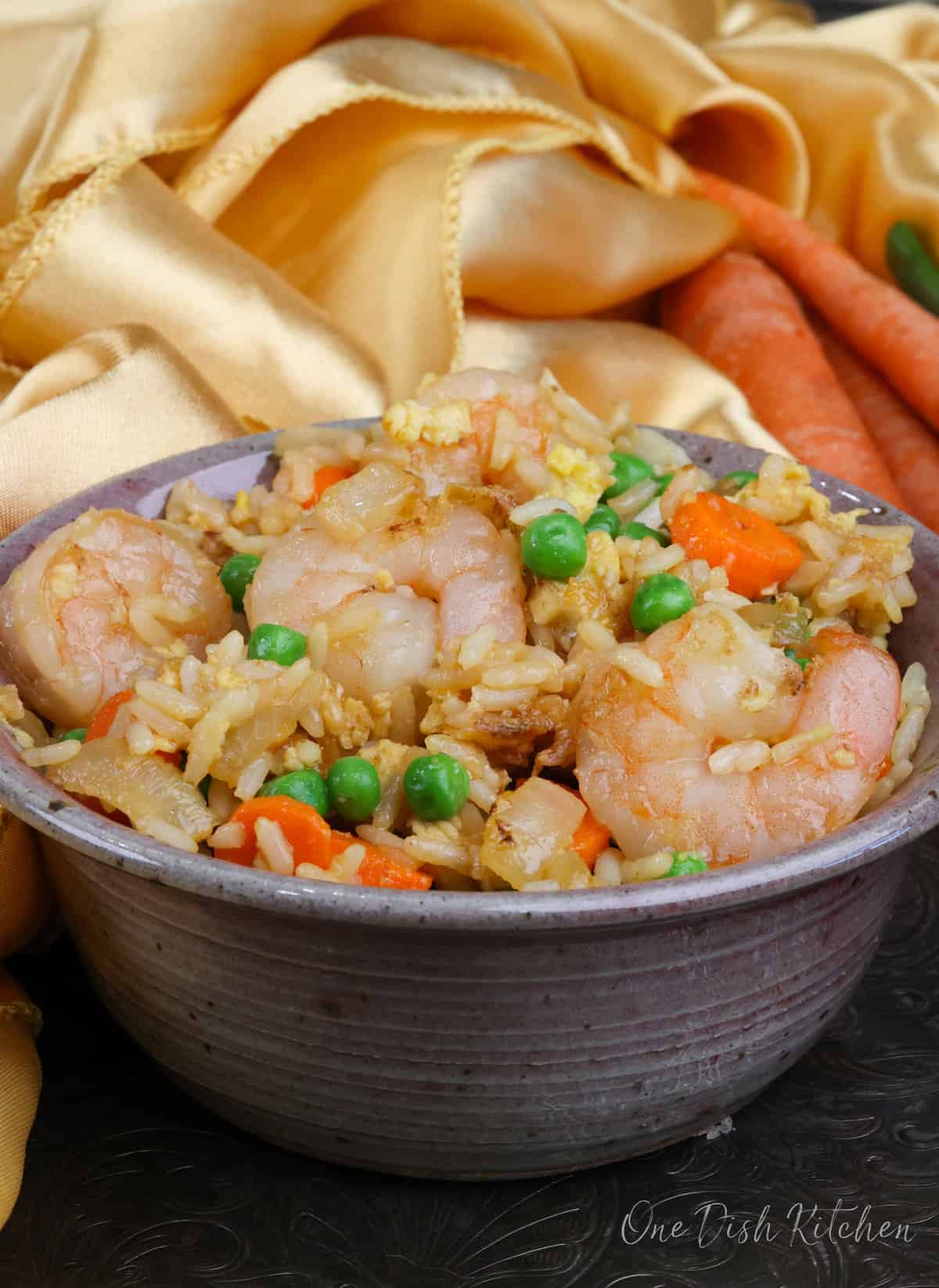 a bowl of shrimp fried rice next to three carrots and a gold napkin