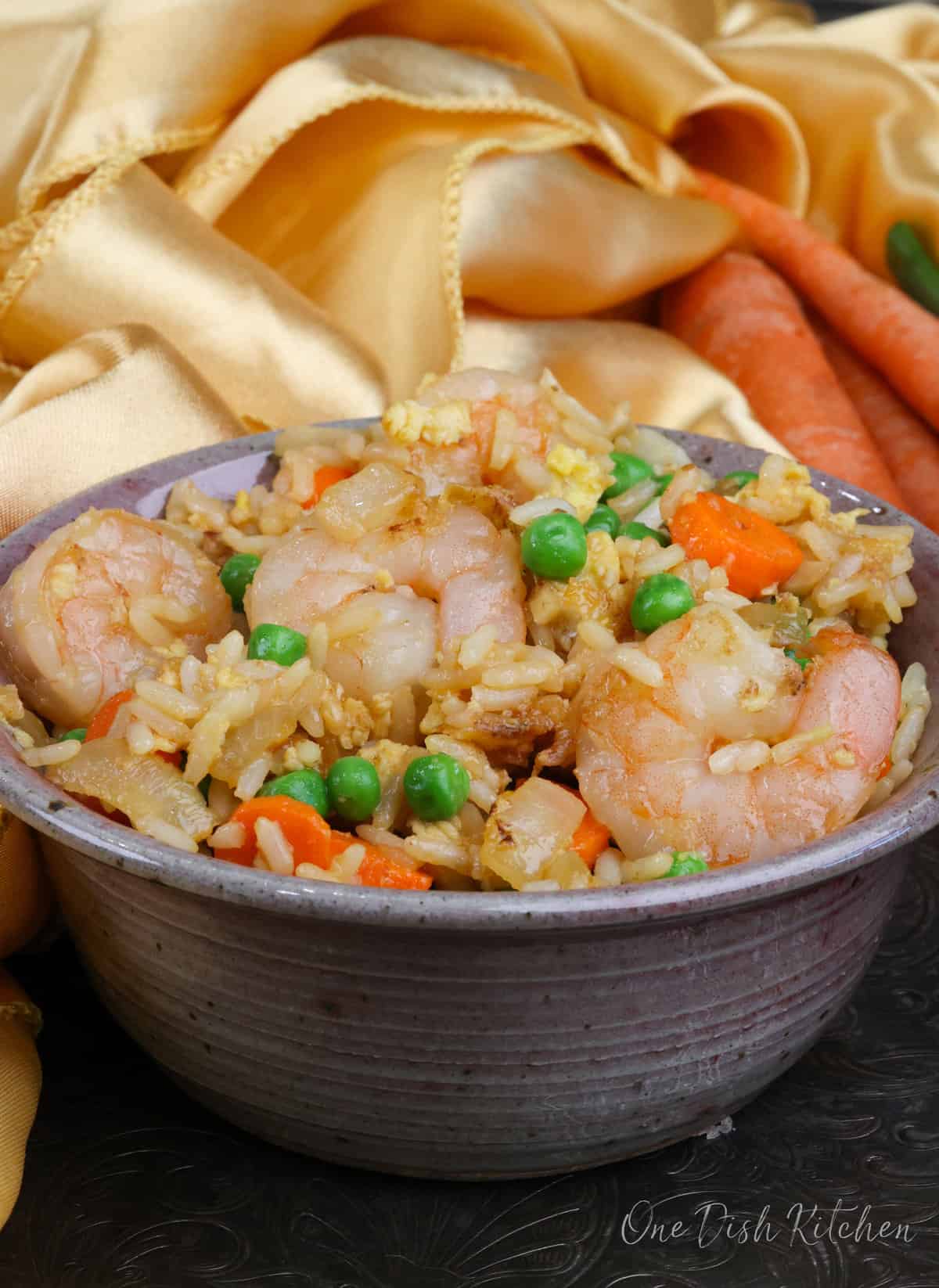 a bowl of shrimp fried rice next to three carrots and a gold napkin.