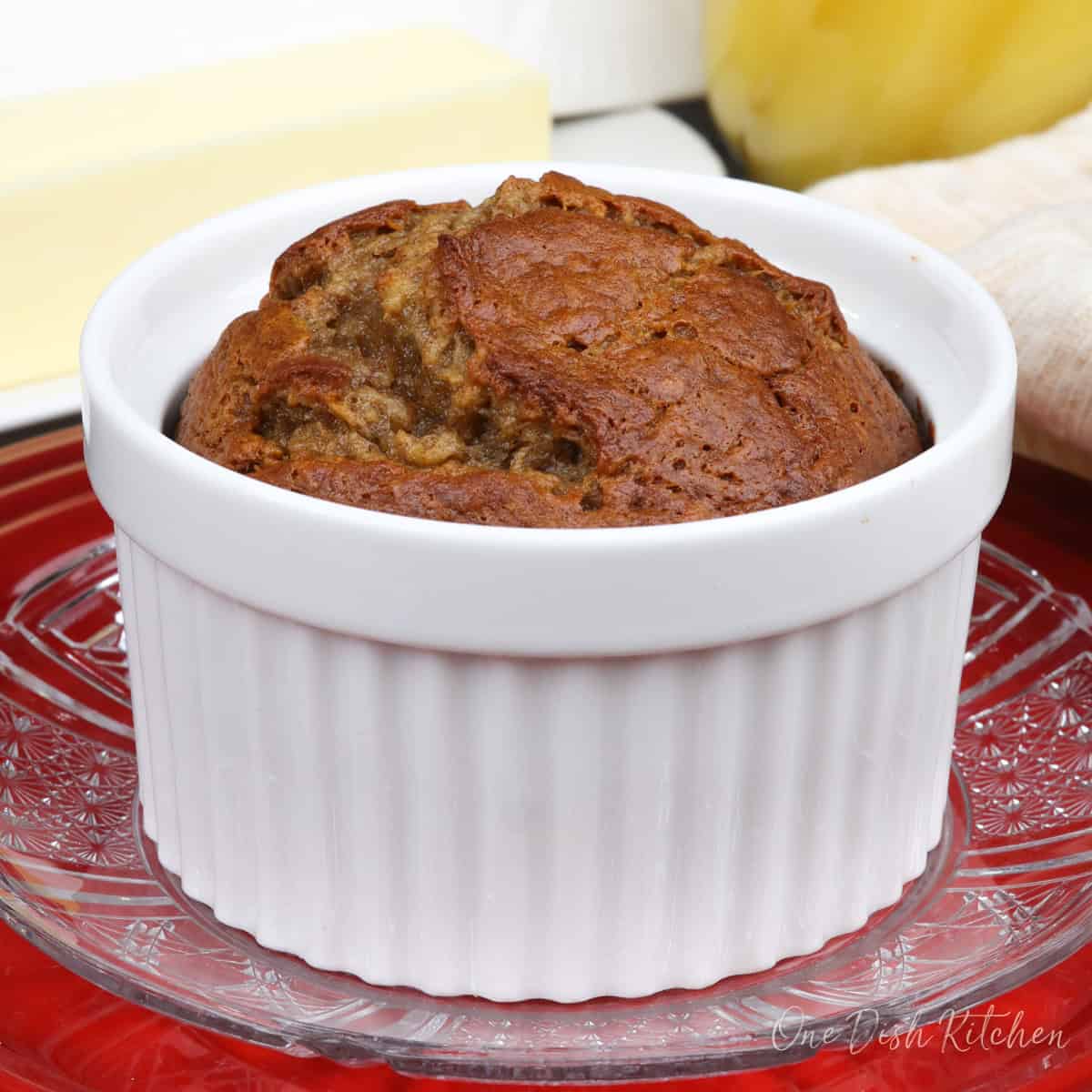 a small banana nut bread baked in a ramekin on a red plate next to a plate of butter and a glass of orange juice.