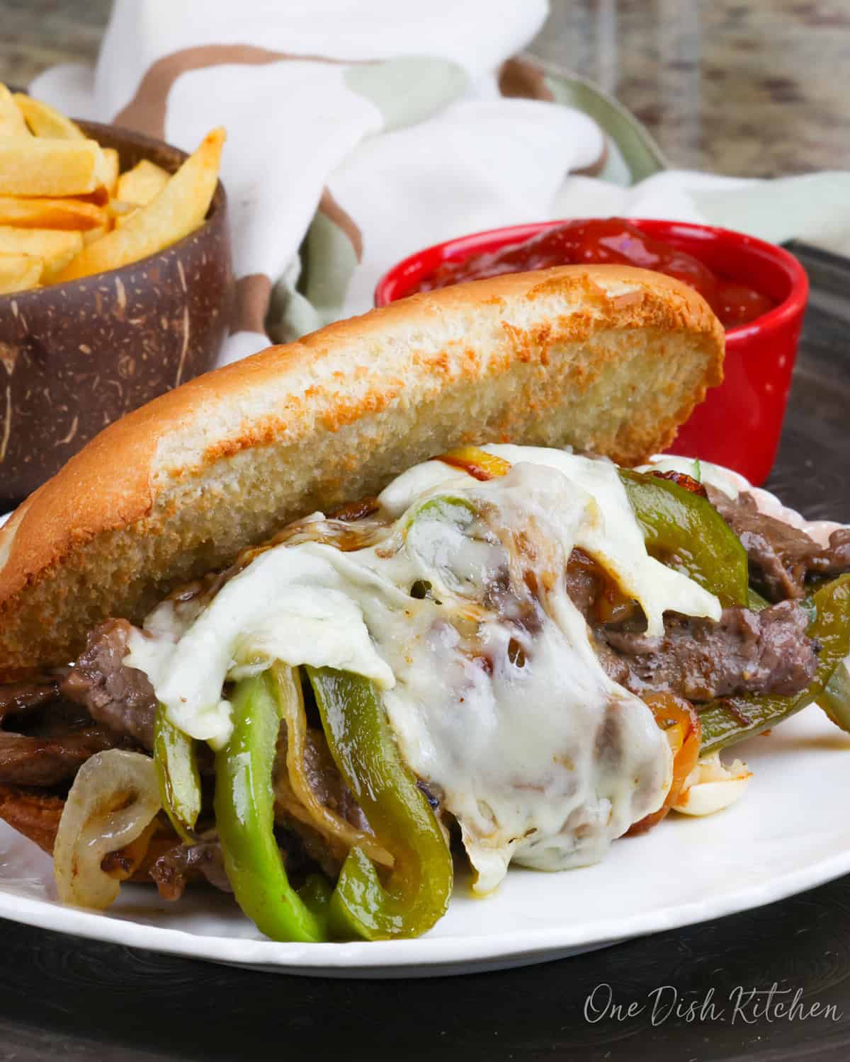 a philly cheesesteak on a white plate next to a bowl of french fries and a red bowl filled with ketchup.