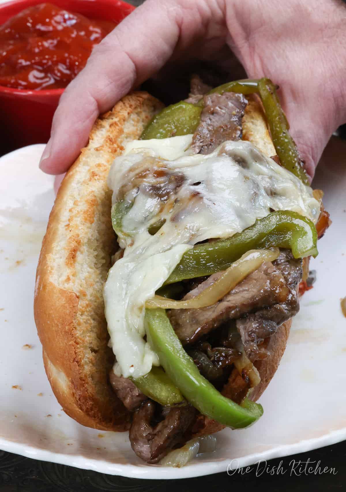 a philly cheesesteak in a mans hand over a plate.