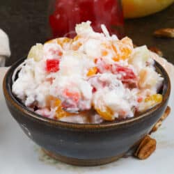 a small bowl of ambrosia salad on a white plate next to pecans, cherries, and a bowl of mixed fruit.