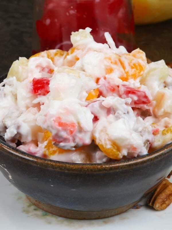 a small bowl of ambrosia salad on a white plate next to pecans, cherries, and a bowl of mixed fruit.