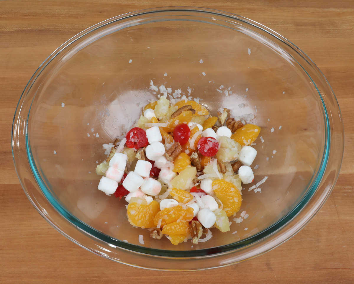 pineapple tidbits, oranges, marshmallows, pecans, and cherries in a mixing bowl.