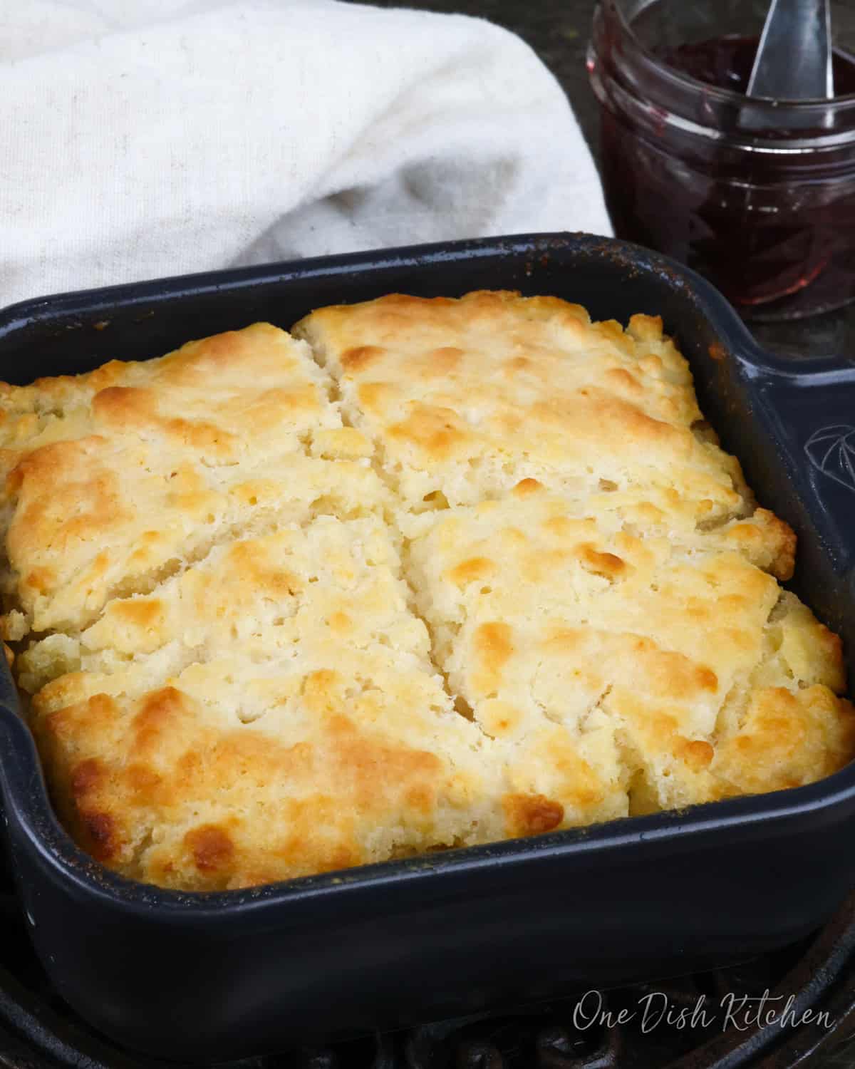 a small square baking dish filled with 4 butter swim biscuits next to a white napkin and a jar of jelly.