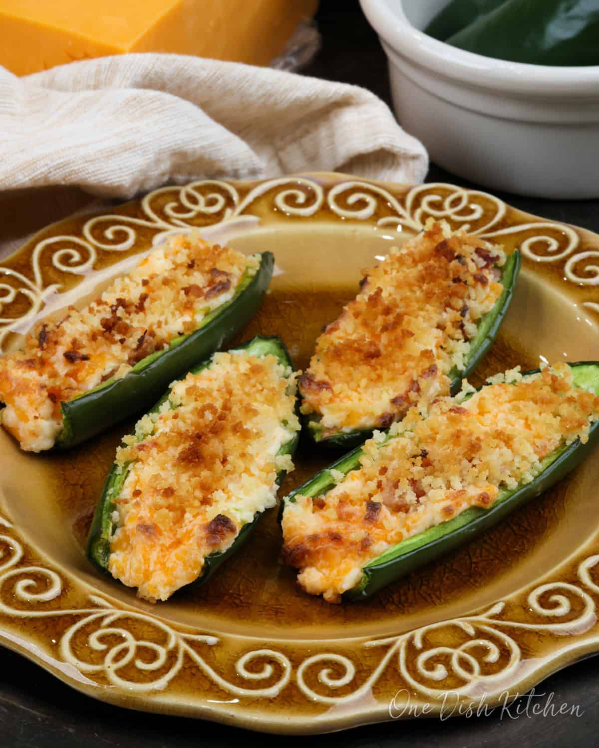 four bacon filled jalapeno poppers on a brown plate next to a bowl of fresh jalapenos and a block of cheddar cheese.