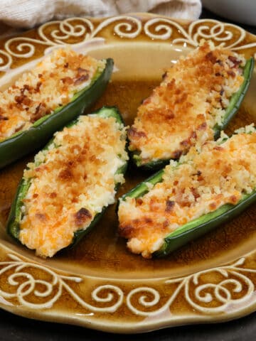 four bacon filled jalapeno poppers on a brown plate next to a bowl of fresh jalapenos and a block of cheddar cheese.