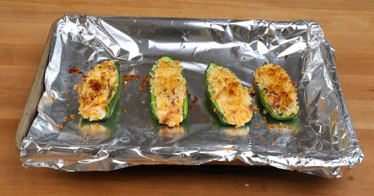 four jalapeno poppers with bacon on a foil lined baking sheet.