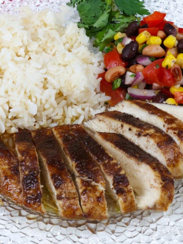 jerk chicken breast on a white plate next to white rice and vegetable salad.