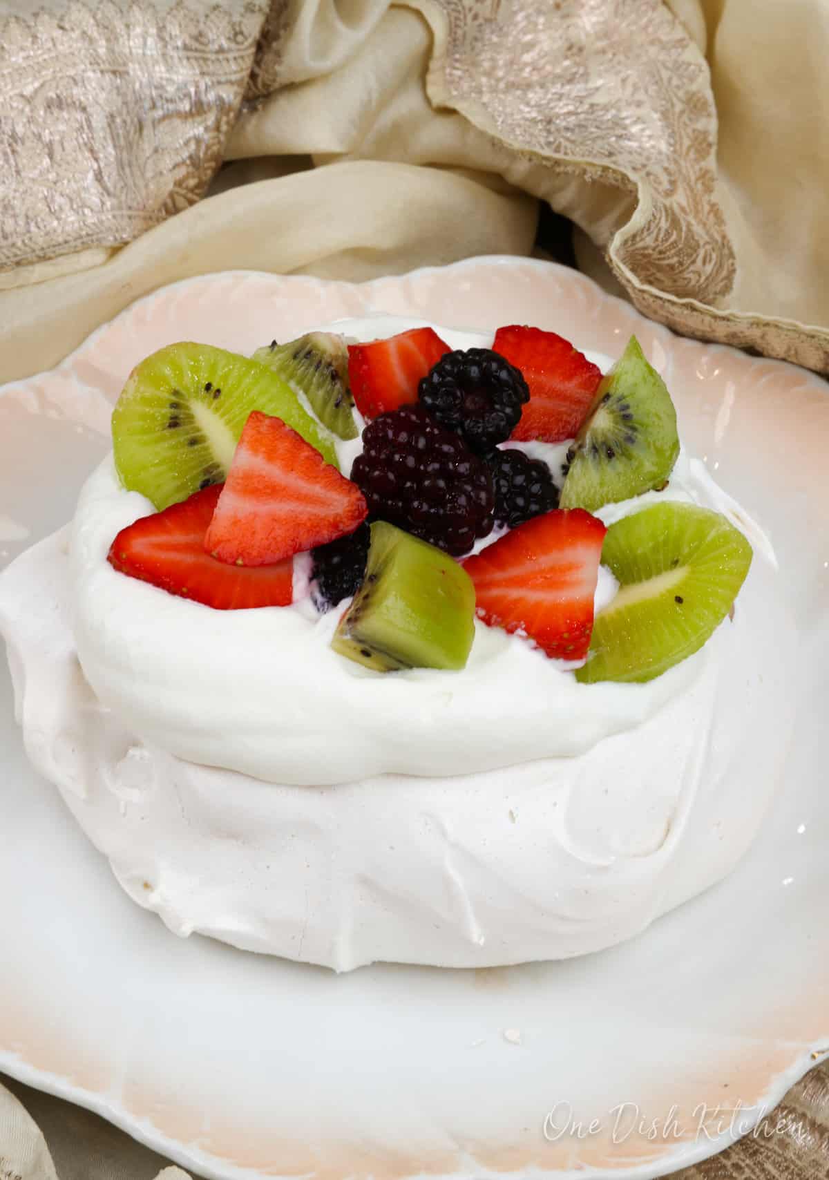 a pavlova on a white plate topped with whipped cream, blackberries, strawberries, and kiwi fruit
