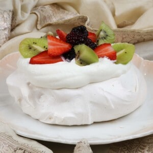 a mini pavlova topped with fresh fruit on a white plate next to a cream napkin