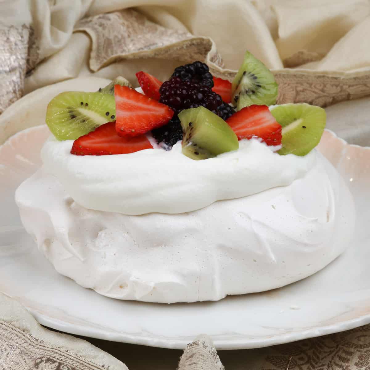 a mini pavlova topped with fresh fruit on a white plate next to a cream napkin.