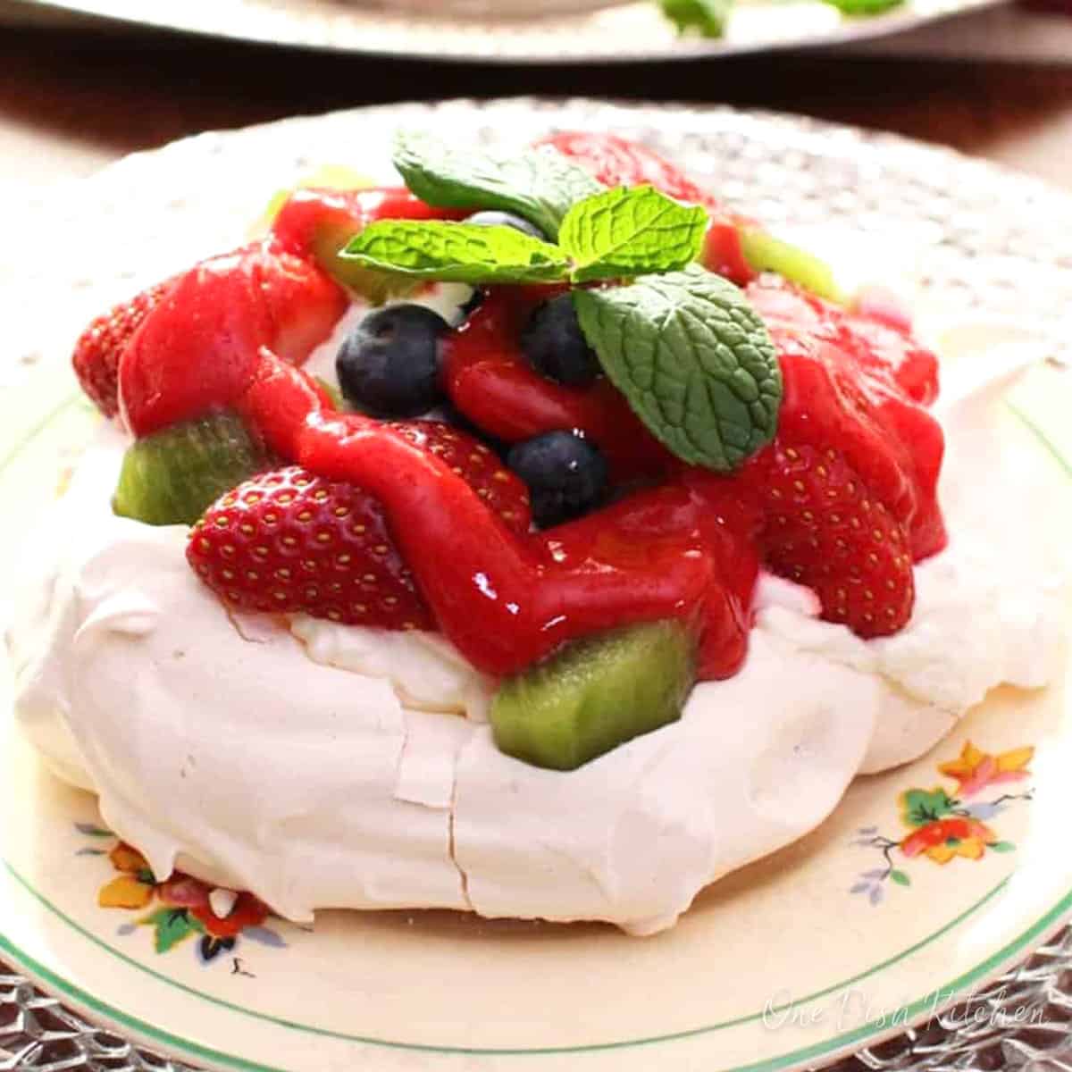 a pavlova topped with whipped cream, coulis, and fresh fruit on a plate.