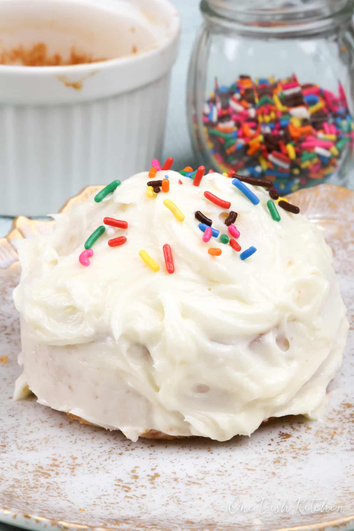 a small white cake on a white plate topped with colored sprinkles next to a ramekin.