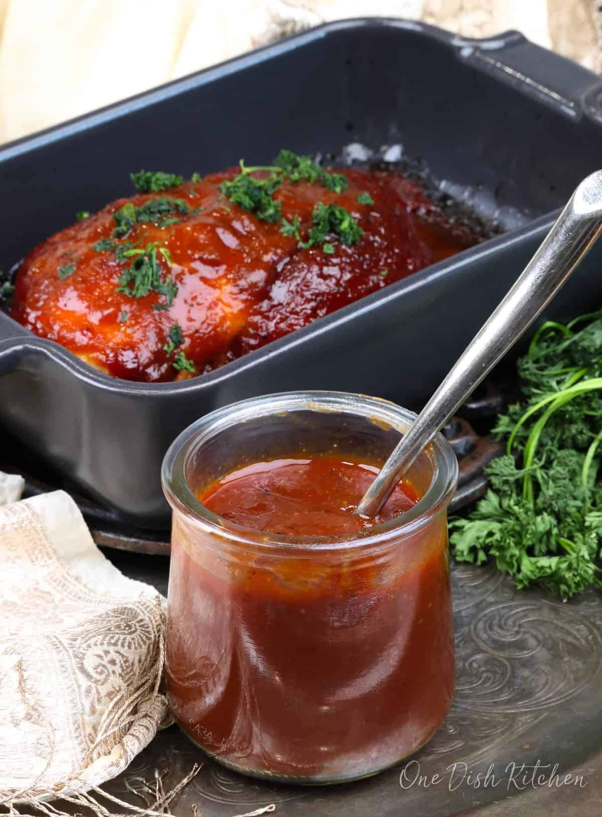 a small jar of bbq sauce next to a chicken breast in a baking dish coated with the sauce