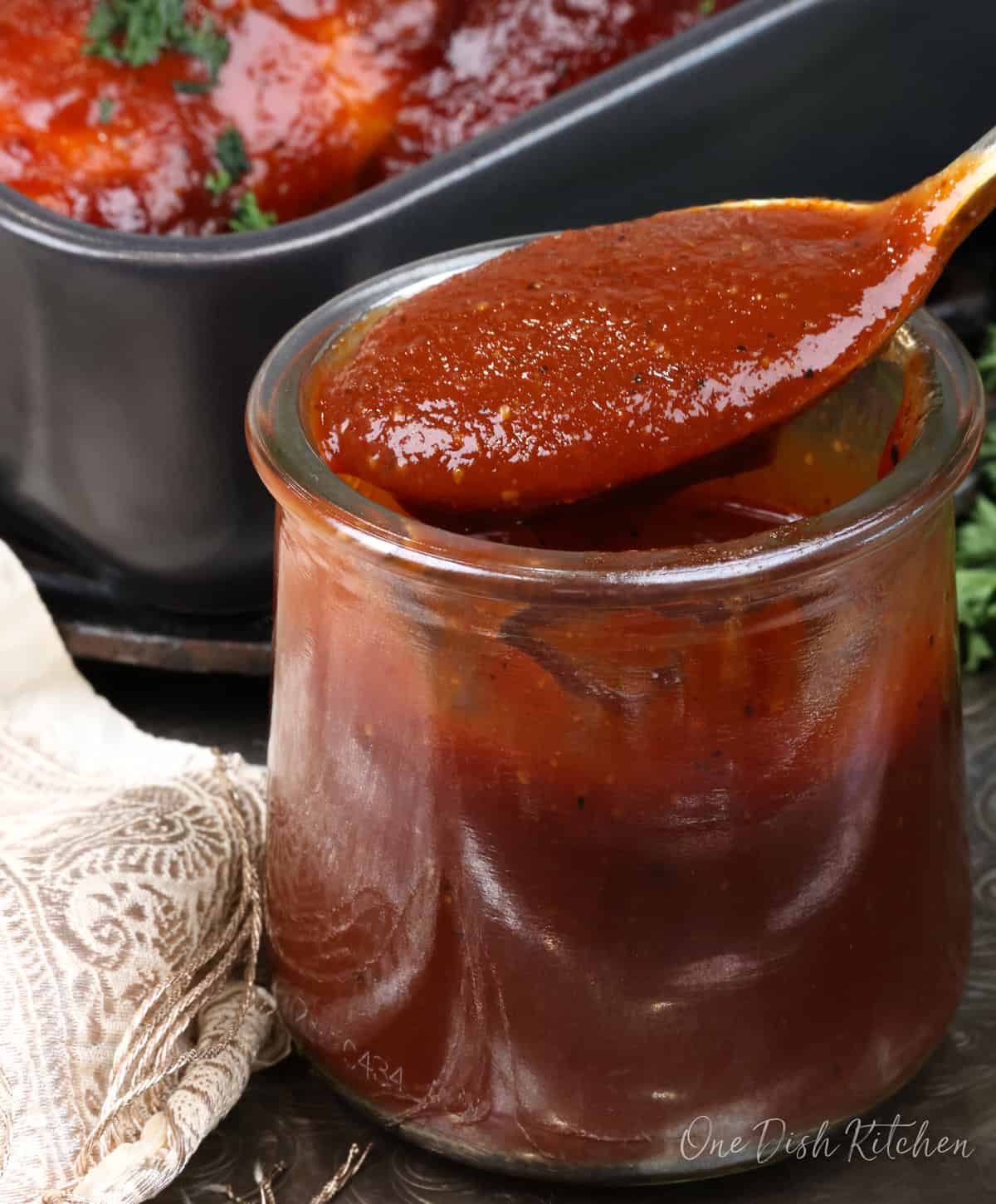 a spoonful of barbecue sauce over a jar with a bbq chicken in the background