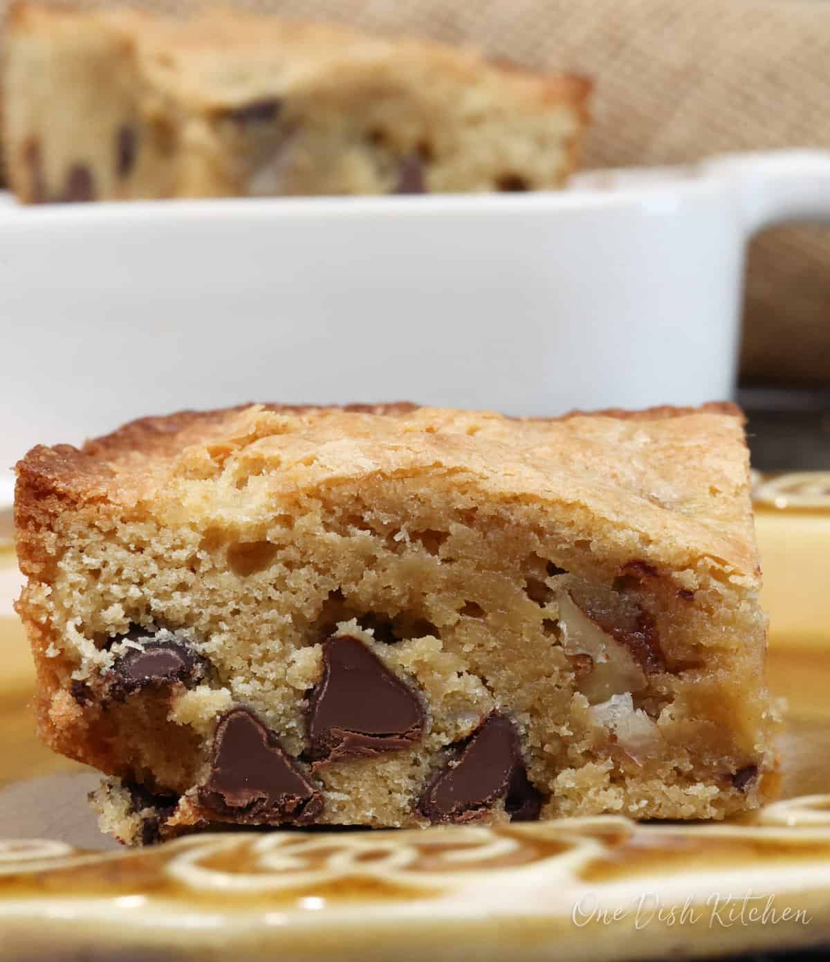 one chocolate chip blondie on a plate next to a bowl of the remaining blondies.