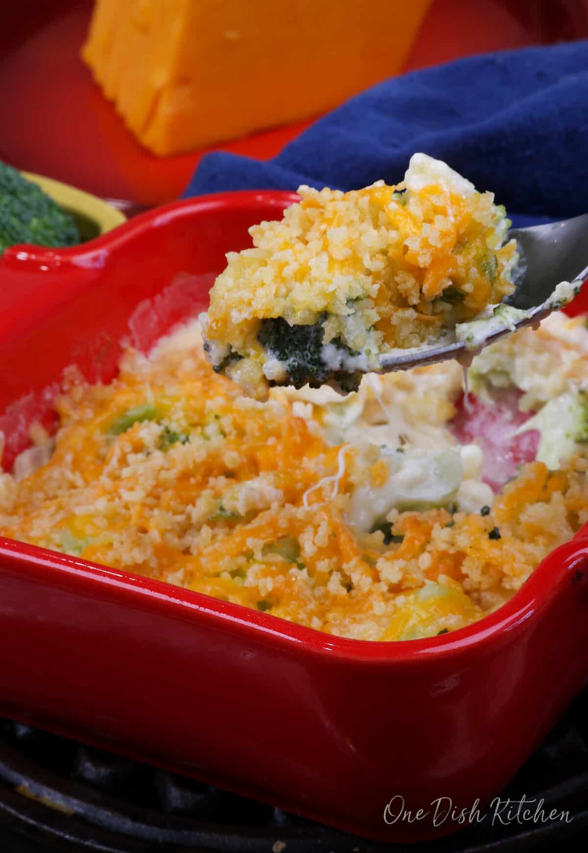 broccoli casserole topped with melted cheddar cheese in a red baking dish with a fork on the side.