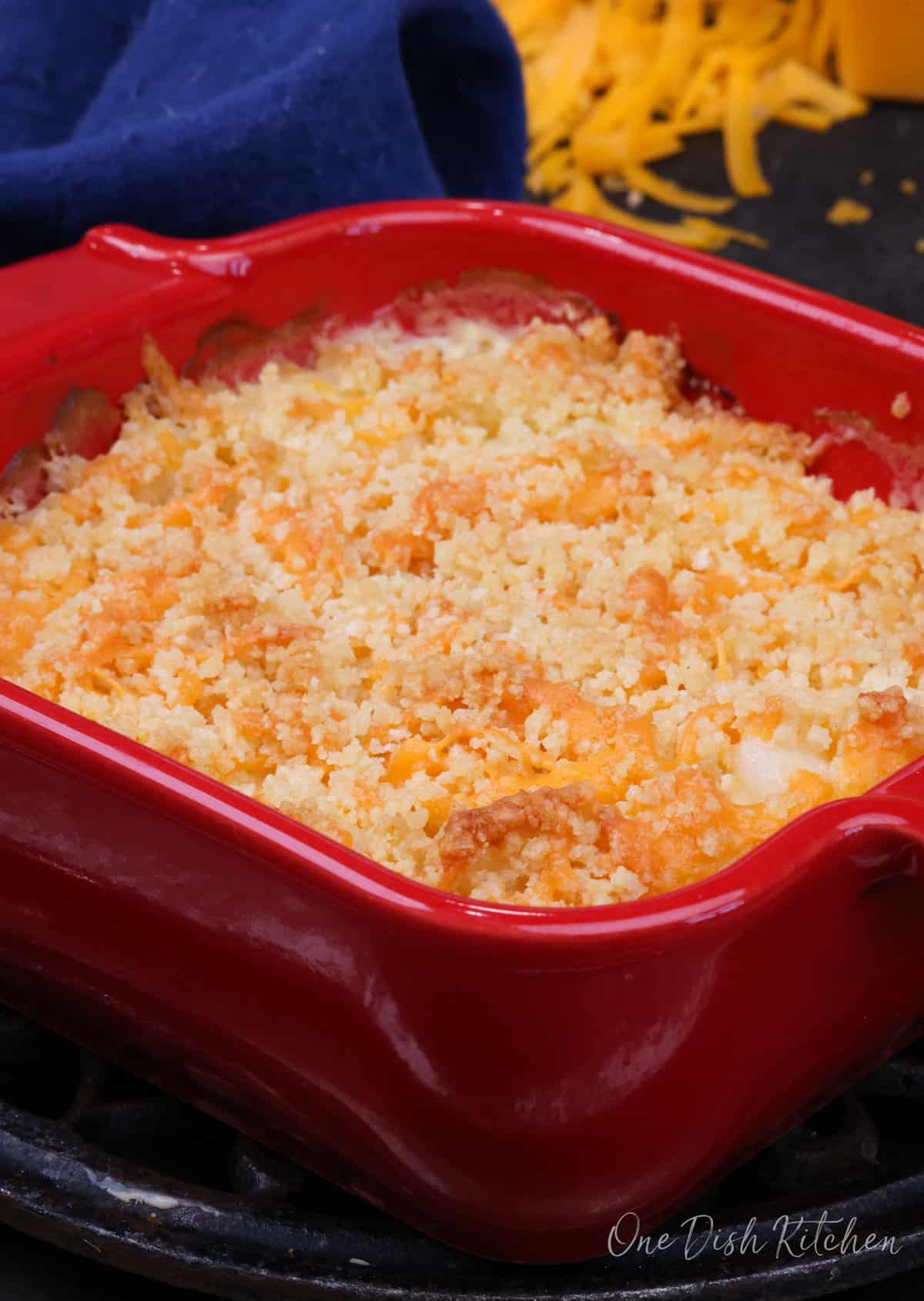 a small hashbrown casserole in a red baking dish next to a blue napkin and a shredded cheddar cheese.