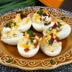 a plate of loaded deviled eggs next to green onions and a wedge of cheddar cheese