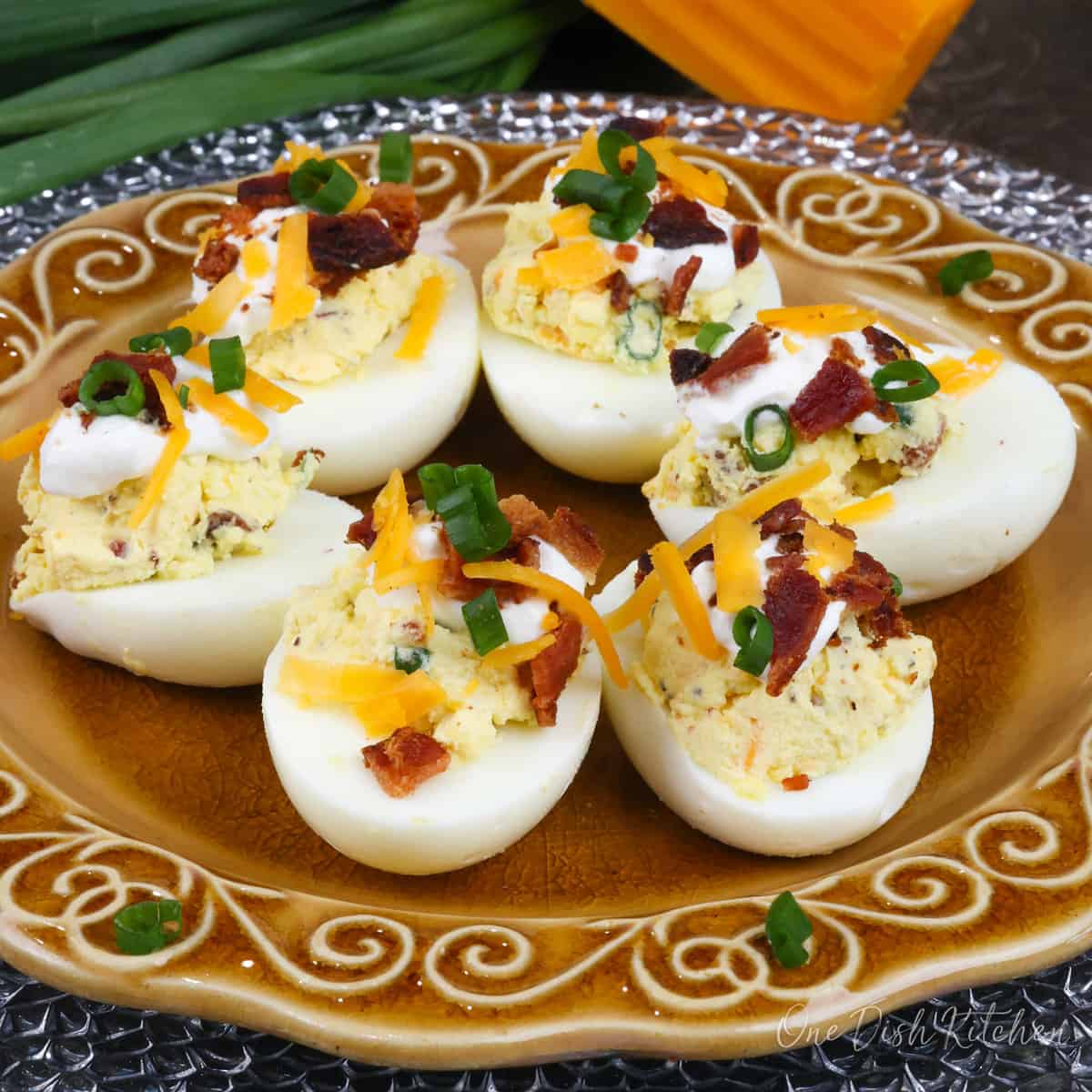 a plate of loaded deviled eggs next to green onions and a wedge of cheddar cheese.