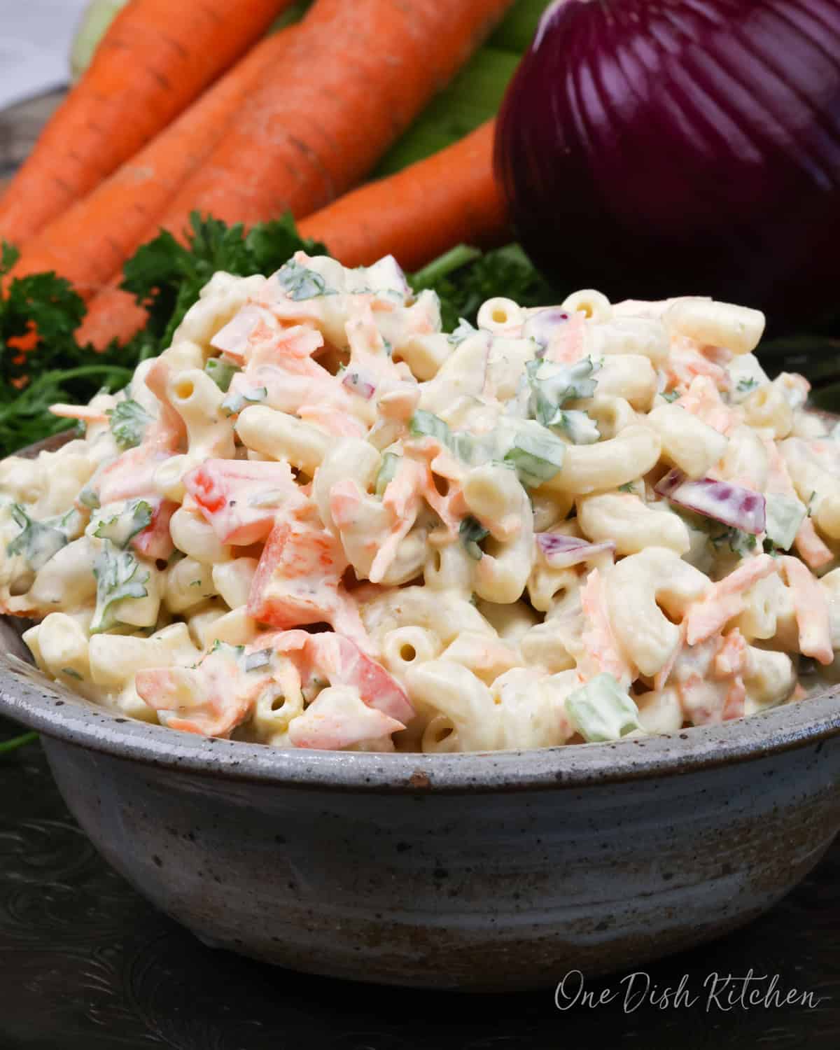 a small bowl of macaroni salad on a silver tray next to a red onion and four carrots.