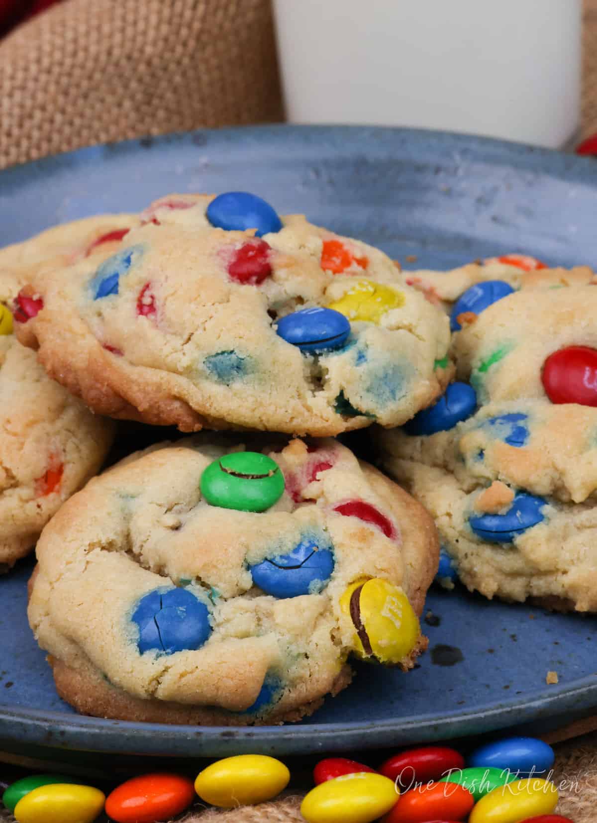 6 m&m cookies on a blue plate next to a glass of milk and colorful m&ms scattered around the plate.