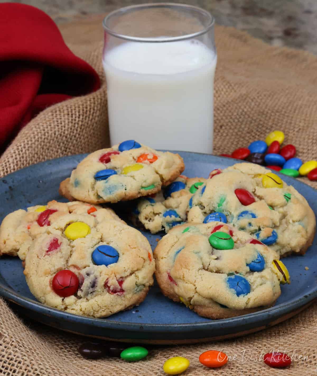 colorful M&M cookies on a blue plate next to a red napkin and a glass of milk.