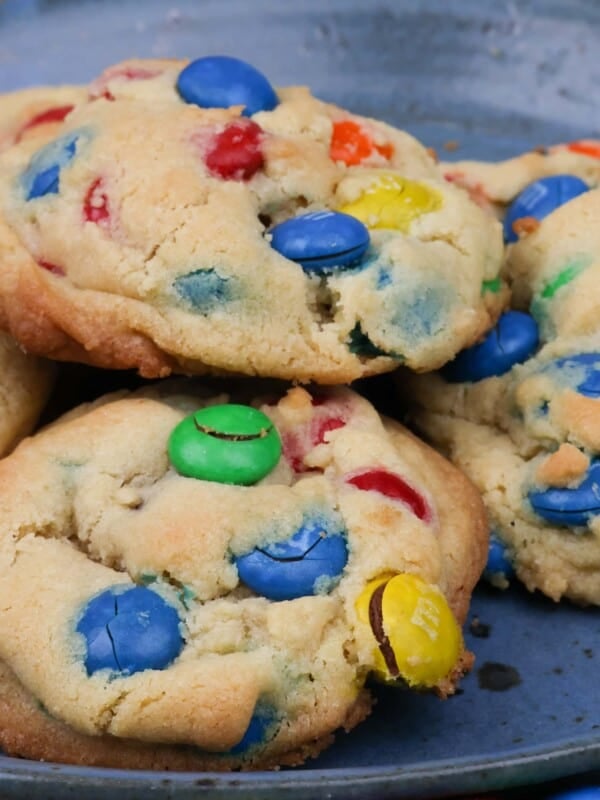 6 m&m cookies on a blue plate next to a glass of milk and colorful m&ms scattered around the plate