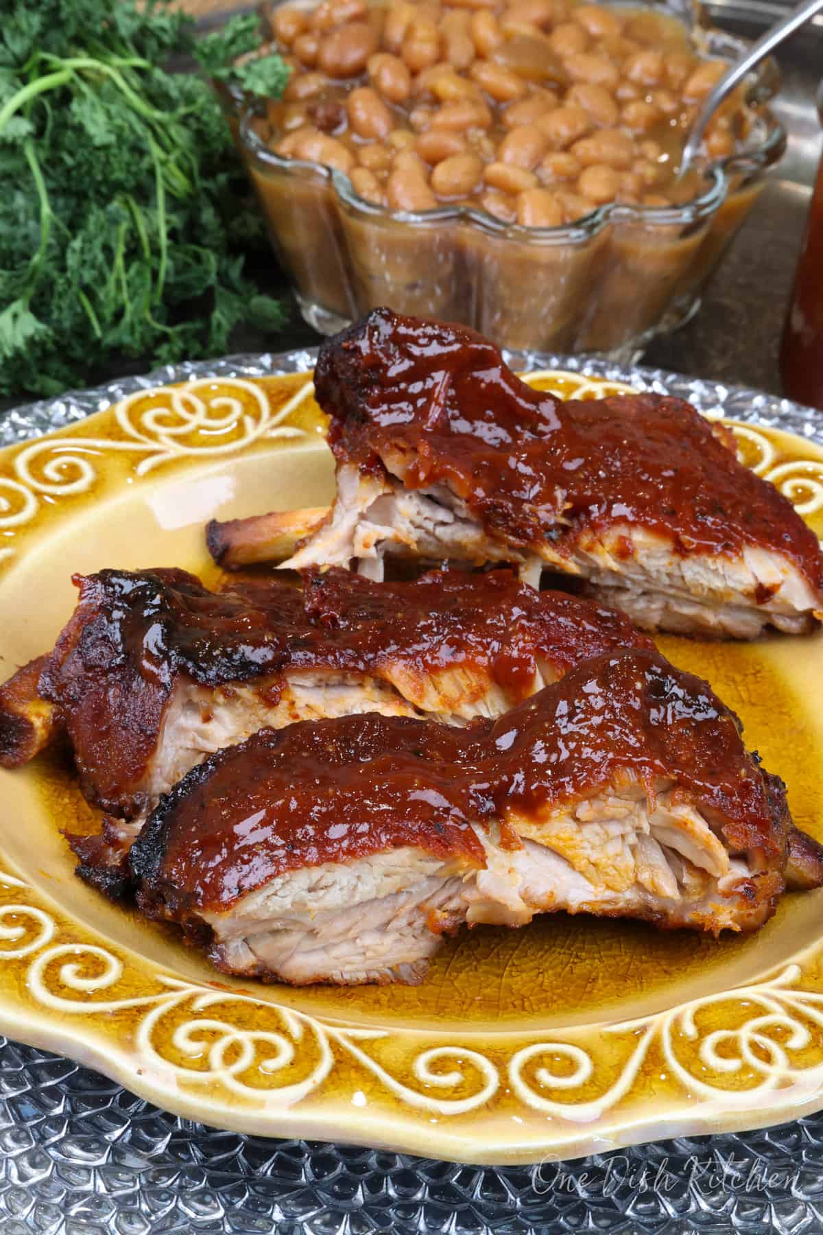 three baby back ribs with BBQ sauce on a gold plate next to a bowl of baked beans.
