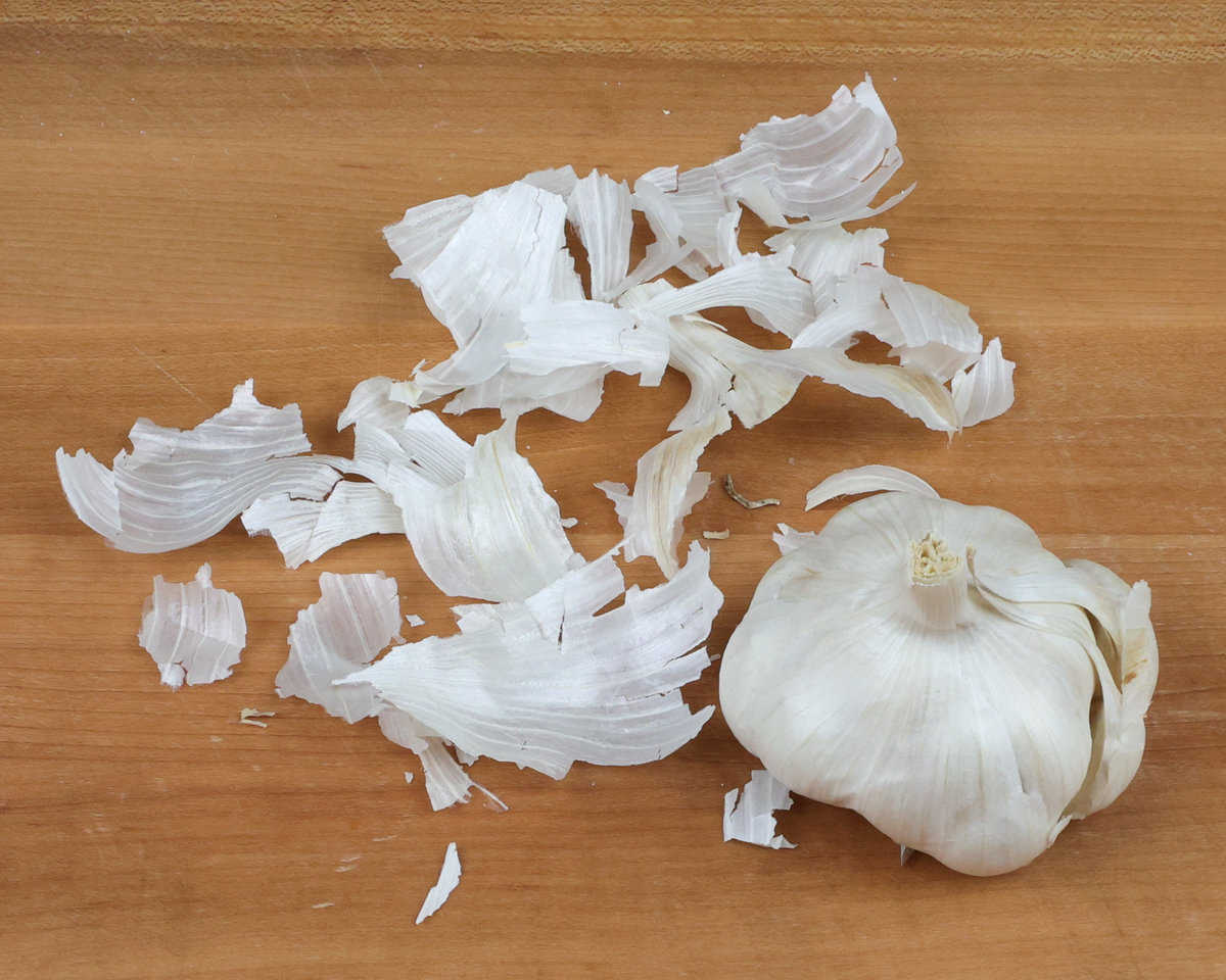 the papery outer layers of a head of garlic resting next to the head of garlic on a cutting board.