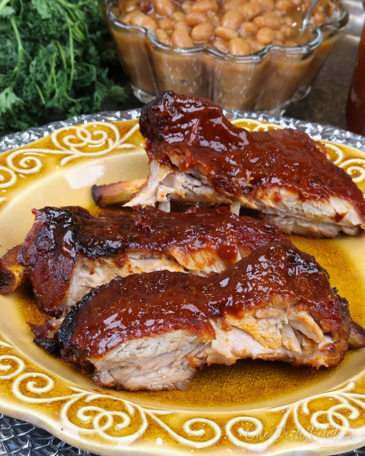 three baby back ribs with BBQ sauce on a gold plate next to a bowl of baked beans.